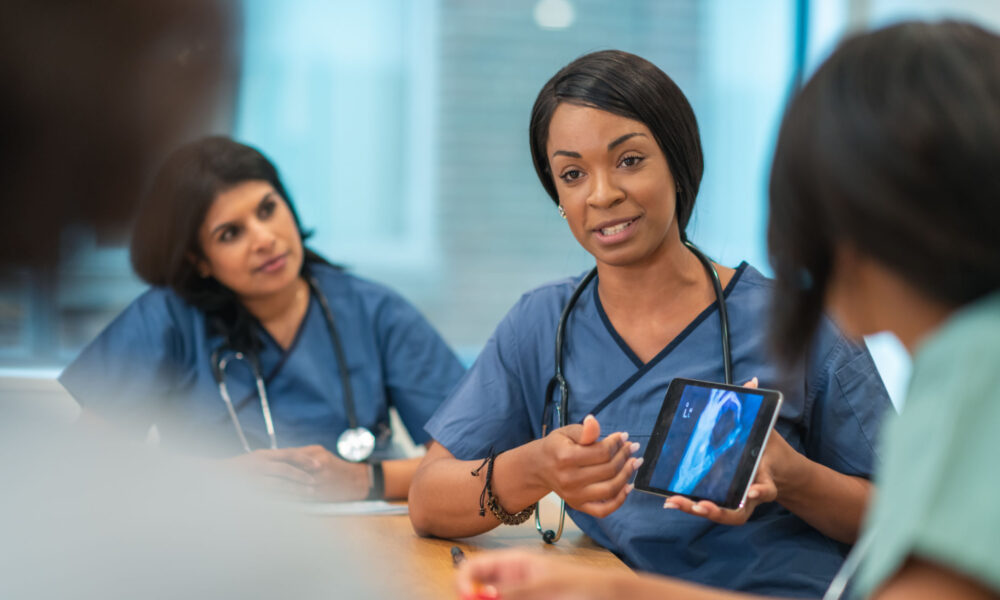 A Black female doctor leads a meeting with her medical team. She is showing the group a medical x-ray on a digital tablet. The multi-ethnic group of medical professionals is seated around a table in a conference room. They are discussing a treatment plan for their patient.
